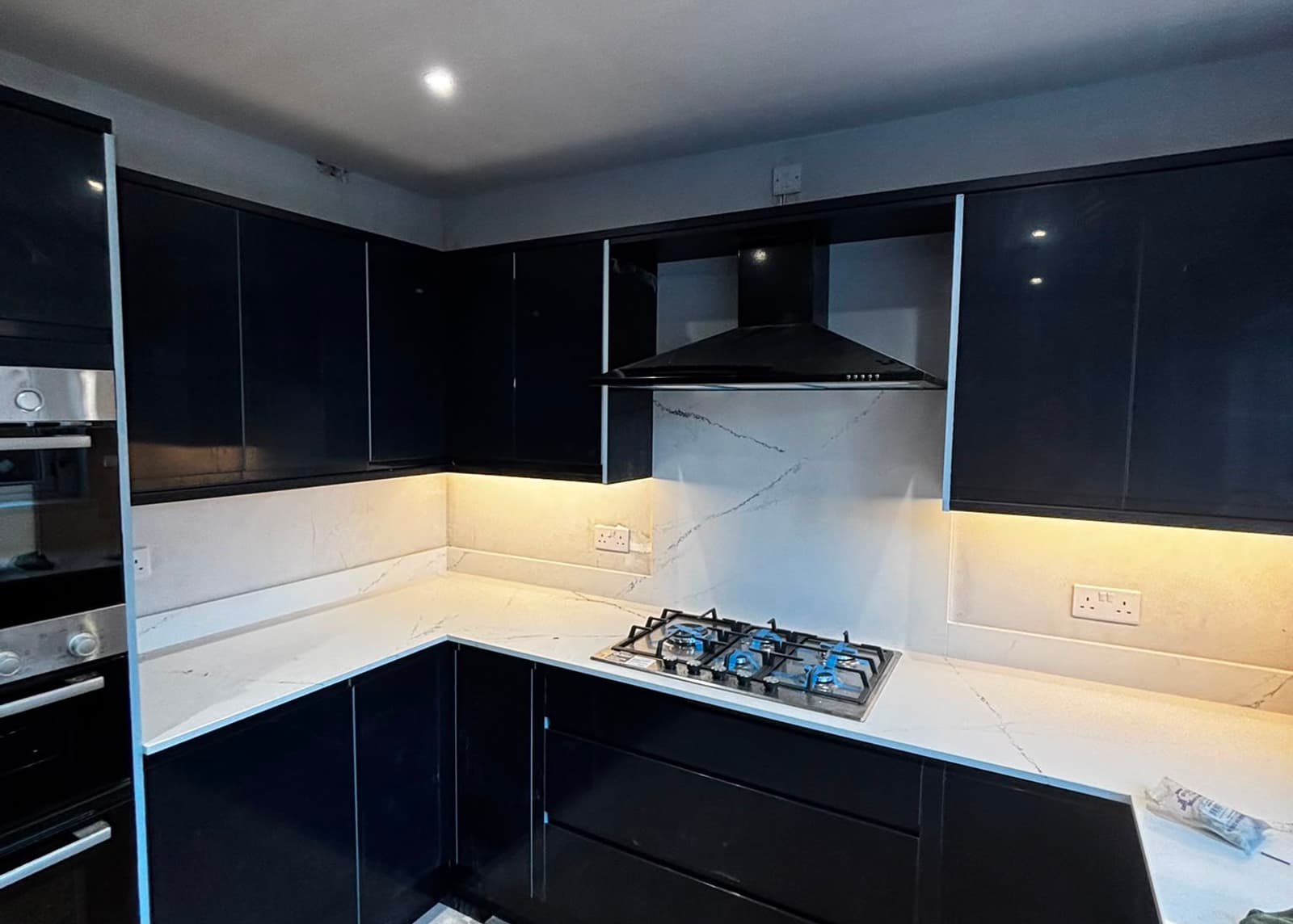 Newly fitted black gloss kitchen with white quartz worktops and lit splashback, by SMM Building Services